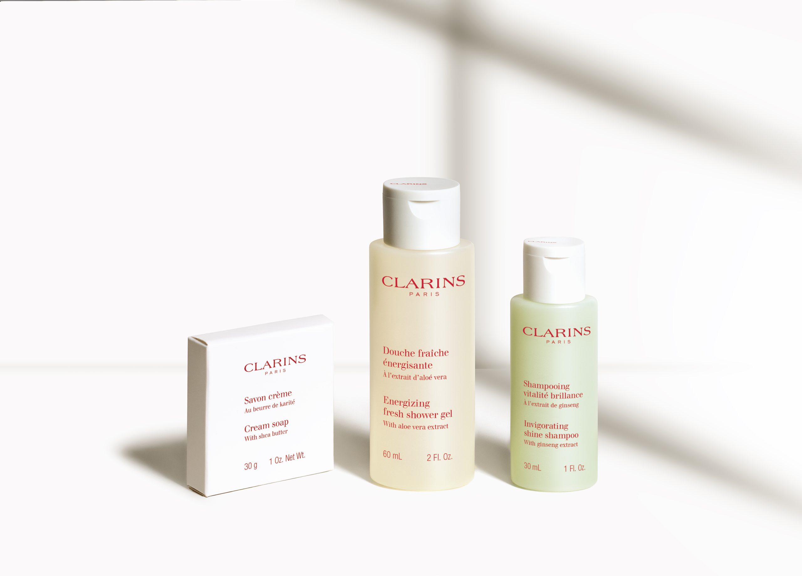 Clarins Toilettenartikel in Reisegröße, darunter Cremeseife, erfrischendes Duschgel und Glanzshampoo, arrangiert auf einem strahlend weißen Hintergrund.