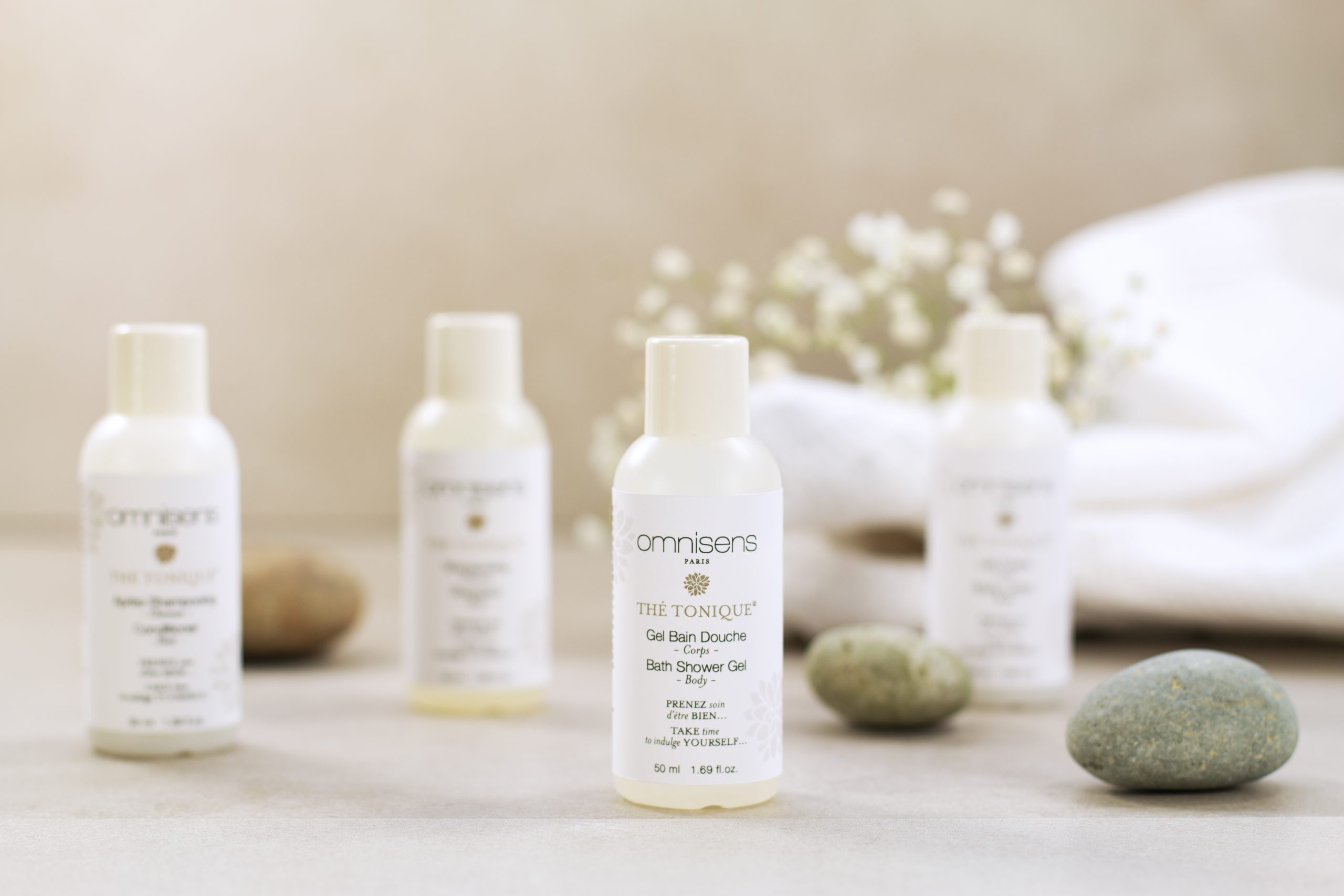 Mini Omnisens Paris „Thé Tonique“ Dusch- und Badegel-Flasche inmitten von Steinen im Spa-Stil und sanft gefalteten Handtüchern.