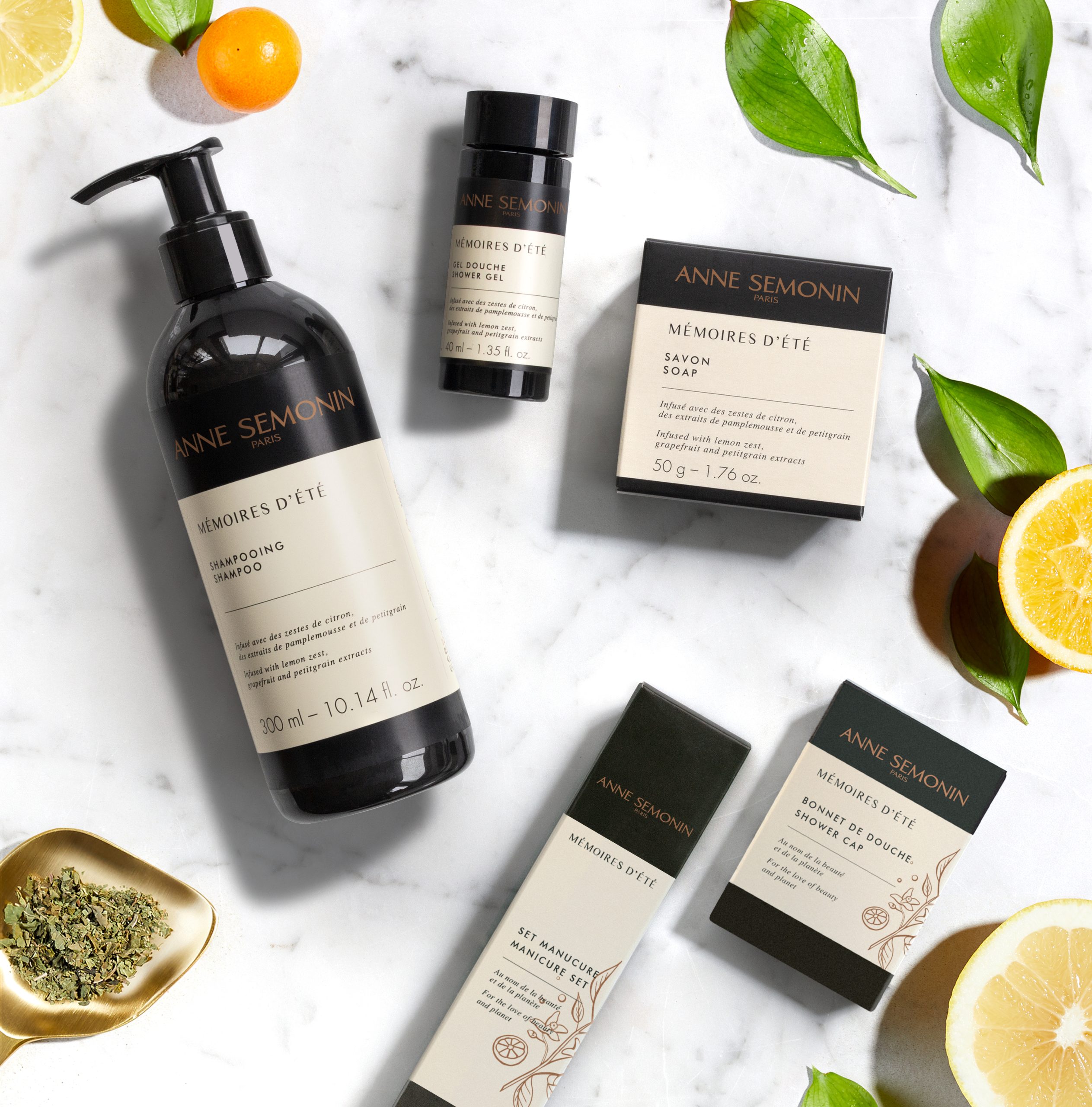 Anne Semonin „Mémoires d’Été“-Shampoo, Seife und Pflegeprodukte liegen flach auf einer Marmorfläche, umgeben von Zitrusfrüchten und grünen Blättern.