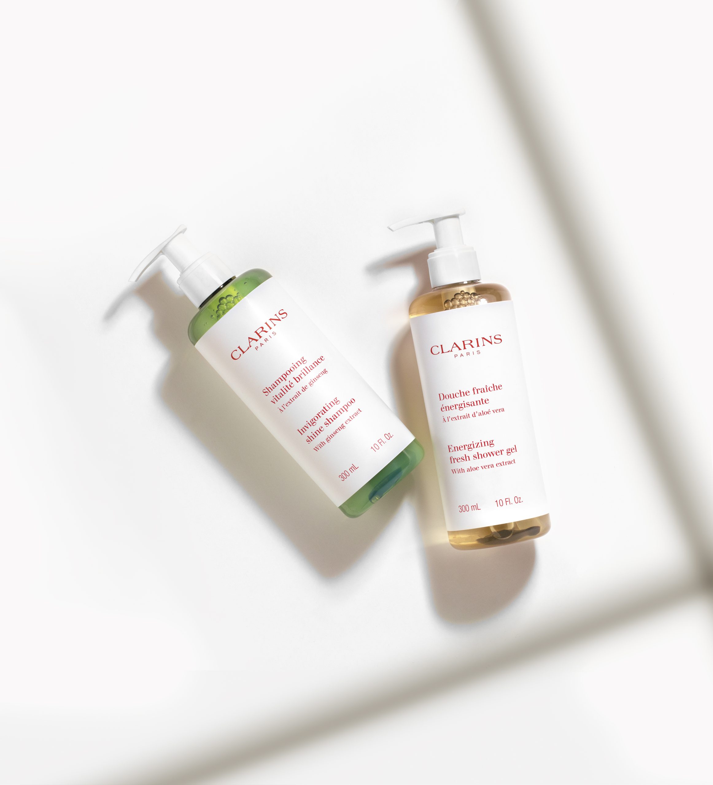 Clarins Flaschen mit dem belebenden Glanz-Shampoo und dem energiespendenden Frische-Duschgel liegen auf einer weißen Fläche im sanften Tageslicht.