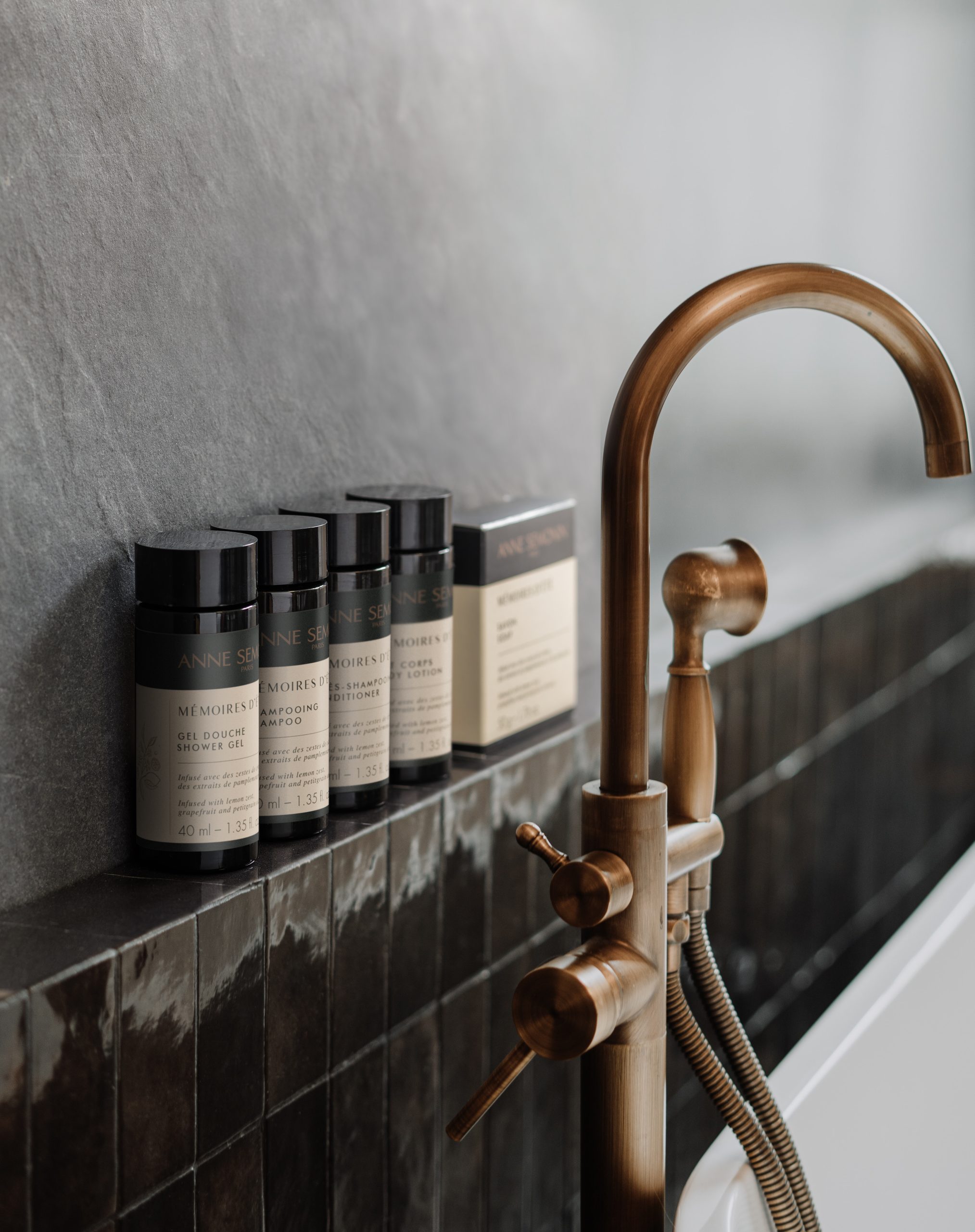 Anne Semonin „Mémoires d’Été“-Mini-Badeprodukte, arrangiert auf einer gefliesten Badewannenablage neben einem Wasserhahn aus gebürstetem Messing.