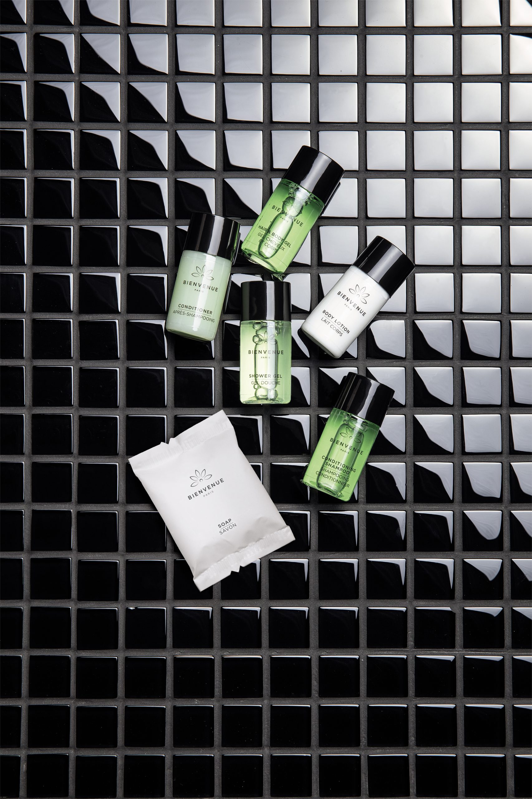 Bienvenue Kleine Toilettenartikel aus Paris, arrangiert auf einer schwarz gefliesten Fläche, darunter Shampoo, Haarspülung, Duschgel, Körperlotion und verpackte Seife.