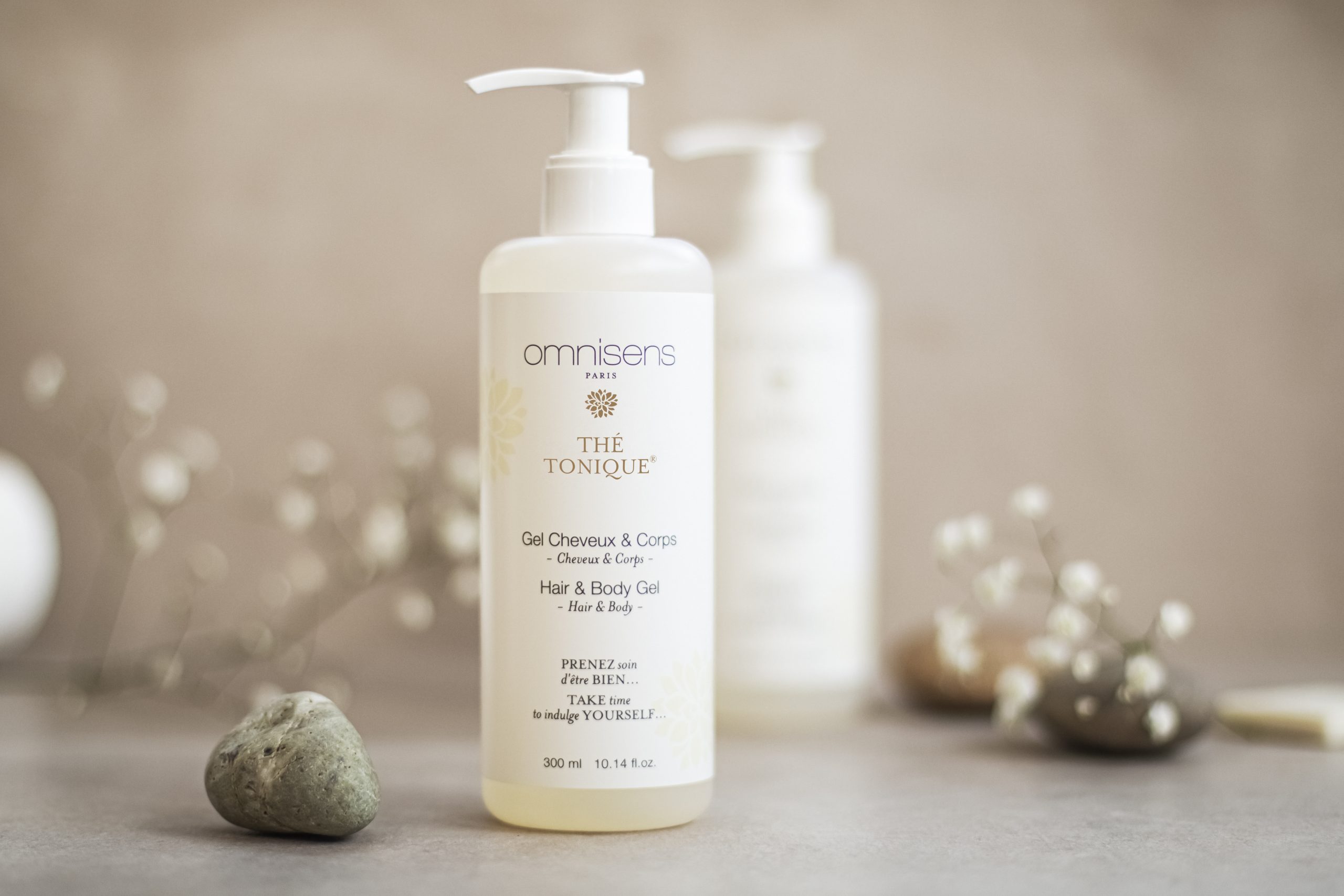 Omnisens Paris „Thé Tonique“ Haar- und Körpergel-Flasche, präsentiert auf einem neutralen Untergrund mit Steinen und zarten floralen Akzenten.