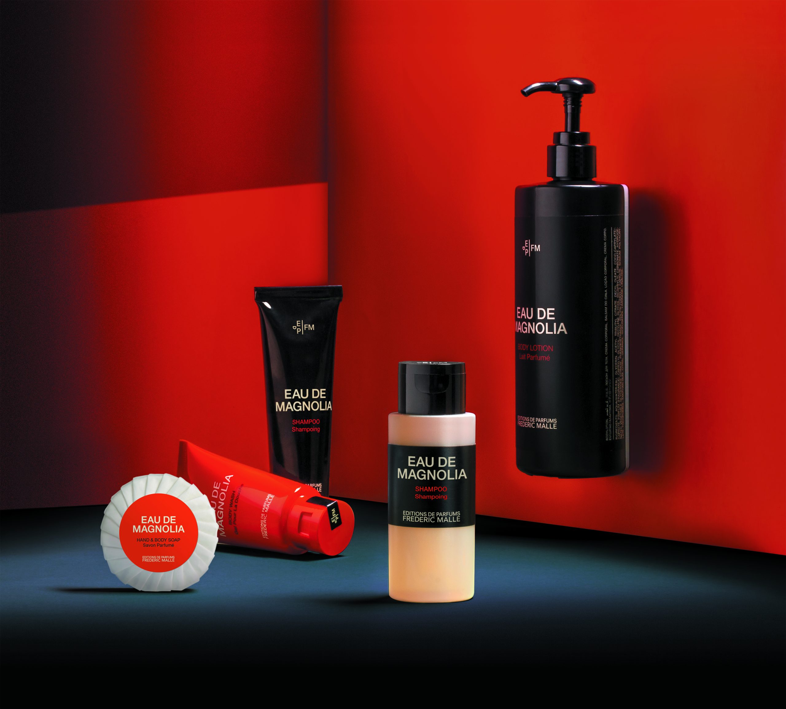 Die Produktpalette von „Eau de Magnolia“ von Editions de Parfums Frédéric Malle wird vor einem kräftigen roten Hintergrund präsentiert, darunter eine Körperlotion mit Pumpe, eine Shampootube, eine Duschgel-Tube und eine rund geformte Seife in Geschenkverpackung, inszeniert mit starker Beleuchtung und tiefen Schatten.