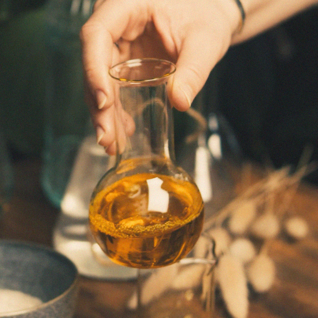 Eine Hand, die eine kleine Glasflasche mit goldfarbener Flüssigkeit hält, die aus natürlichen Inhaltsstoffen besteht, vor einem sanften, in warmen Farbtönen gehaltenen Hintergrund, der an ein Kosmetik- oder Hautpflegeprodukt erinnert.