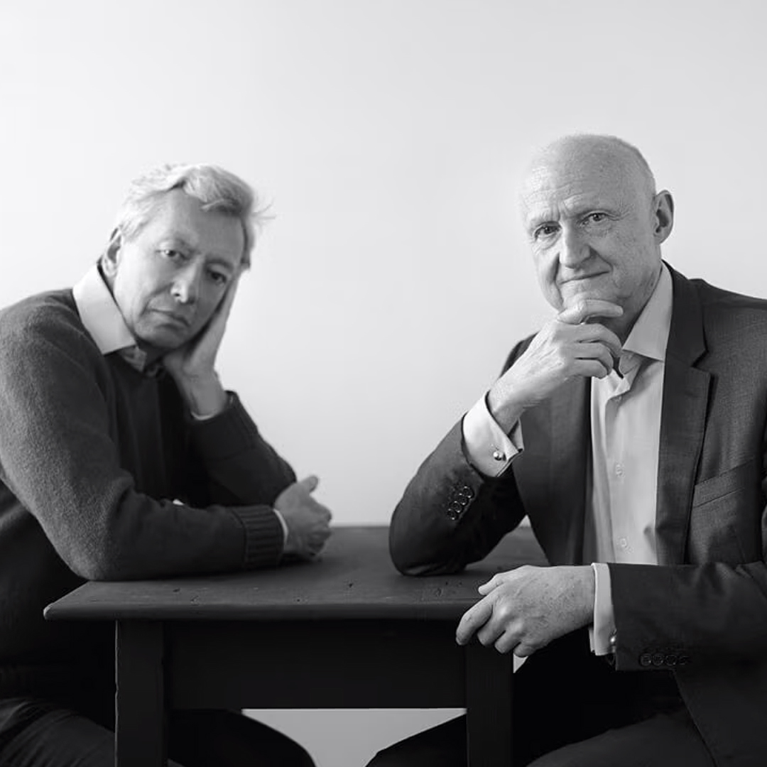 Schwarz-Weiß-Porträt von Frédéric Malle und Dominique Ropion, die an einem Tisch sitzen und in ein minimalistisches Studio mit sanfter Beleuchtung und neutralem Hintergrund in die Kamera blicken.