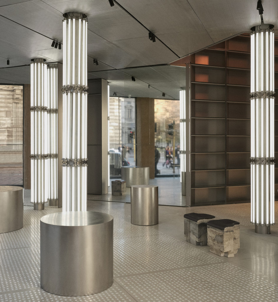 Moderne Laden- oder Ausstellungsraumgestaltung mit metallischen, zylindrischen Ausstellungselementen, hohen, vertikalen Leuchten und reflektierenden Oberflächen, die einen eleganten und zeitgemäßen architektonischen Raum schaffen.