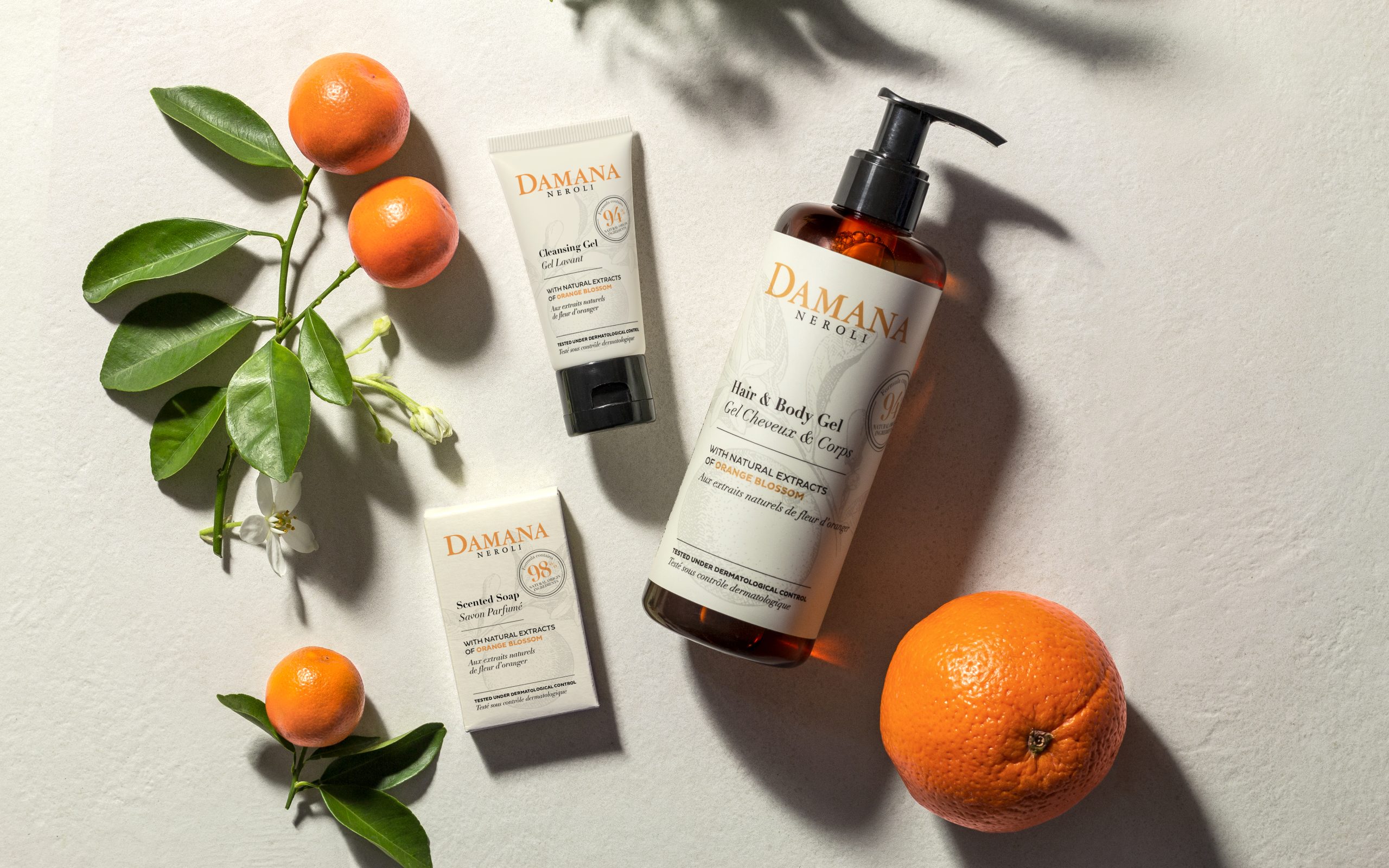Damana Neroli Hautpflegeprodukte, präsentiert auf einem hellen Untergrund neben frischen Orangen und grünen Blättern, darunter eine Flasche Haar- und Körpergel, eine Tube Reinigungsgel und duftende Seife, die die natürlichen Inhaltsstoffe der Orangenblüte und eine frische, von Zitrusfrüchten inspirierte Ästhetik hervorheben.