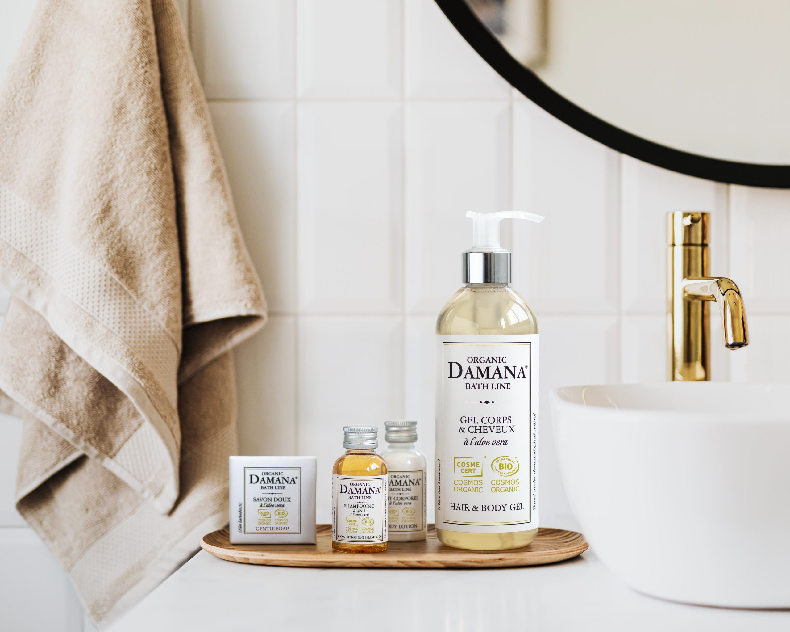 Bio-Badezusätze der Marke Damana, angeordnet auf einem Holztablett neben einem Waschbecken, darunter eine große Flasche Haar- und Körpergel, kleine Shampoo- und Lotionsflaschen sowie eine Seife in einer Schachtel, im Hintergrund weiche Handtücher und eine moderne Badezimmerkulisse.