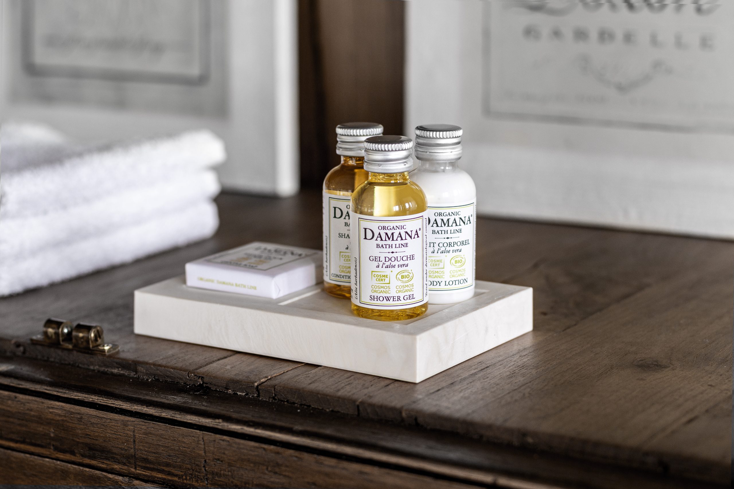 Auf einem weißen Tablett auf einer Holzunterlage sind Bio-Badezusätze der Marke Damana ausgestellt, darunter kleine Flaschen mit Duschgel, Shampoo und Körperlotion sowie eine Seife in einer Schachtel, im Hintergrund liegen gefaltete Handtücher.