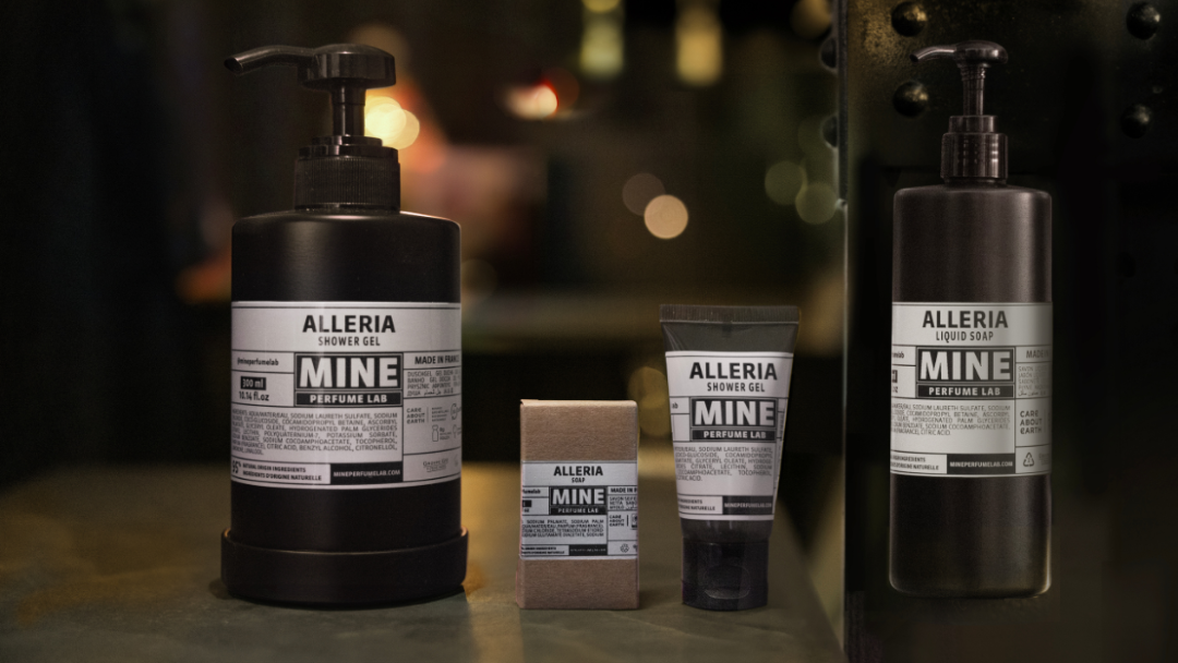 Alleria Mine Perfume Lab ürünleri serisi, loş bir ortamda sergileniyor; seride büyük bir duş jeli şişesi, sıvı sabun pompası, küçük bir tüp ve kutulu sabun yer alıyor. Sıcak bokeh ışıkları, rahat ve zarif bir atmosfer yaratıyor.