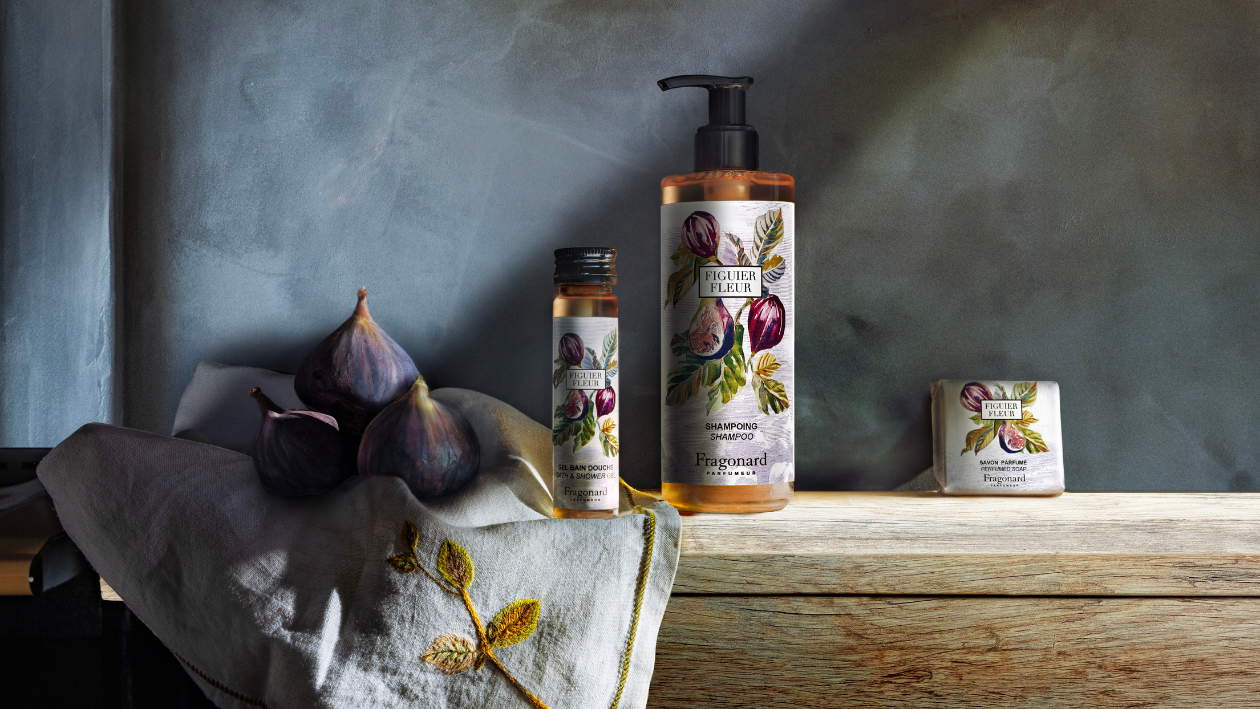 Fragonard Figuer Fleur banyo ürünleri, taze incir ve kumaşlarla süslenmiş ahşap bir yüzey üzerinde sergileniyor; ürün yelpazesi duş jeli şişesi, şampuan pompası ve sabun kalıbını içeriyor ve yumuşak, doğal ışıklandırmanın sağladığı dokulu bir arka planın önünde yer alıyor.