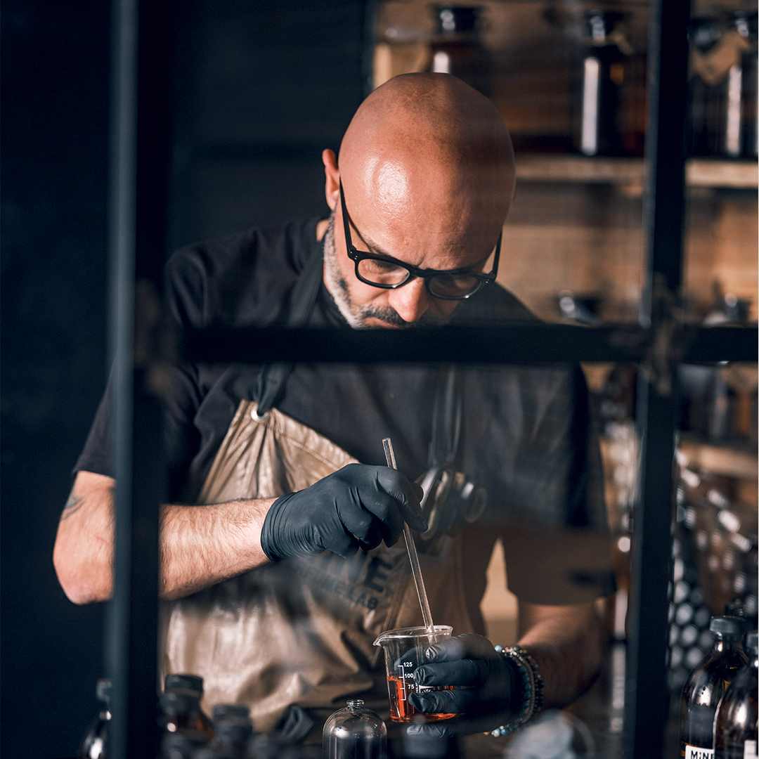 Close-up of Massimo Annunziata in a laboratory setting mixing a liquid in a glass beaker using a pipette, surrounded by small bottles and tools, highlighting a fragrance or cosmetic formulation process with warm, moody lighting.