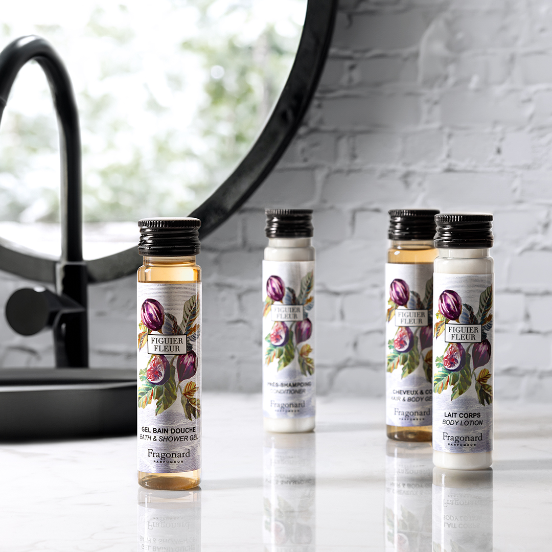 Fragonard Figuer Fleur-Pflegeprodukte in Reisegröße, arrangiert auf einer Marmorfläche neben einem Waschbecken und einem runden Spiegel, bestehend aus kleinen Flaschen mit Duschgel, Shampoo, Haarspülung und Körperlotion mit illustrierten Feigenetiketten.