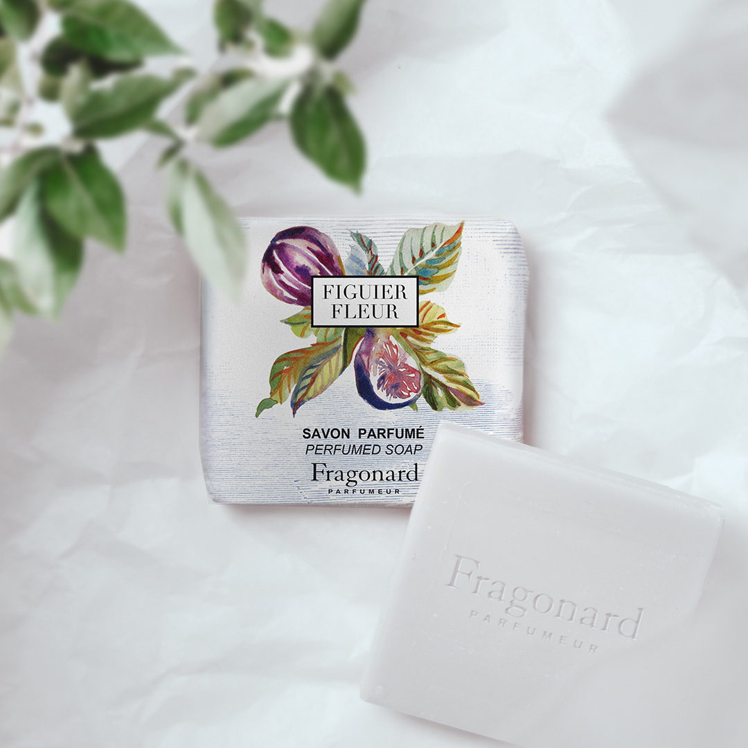 Die parfümierte Seife „Figuer Fleur“ von Fragonard in ihrer illustrierten Verpackung auf einer weichen weißen Unterlage, daneben ein unverpacktes weißes Seifenstück mit dem eingeprägten Fragonard-Schriftzug, im Vordergrund sanft verschwommene Blätter.