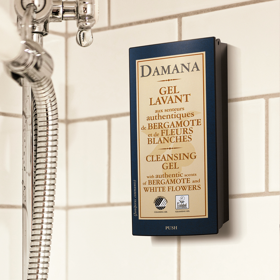 Ein an der Wand montierter Damana-Spender für Reinigungsgel in einer gefliesten Dusche, daneben ein Duschschlauch aus Metall und ein gut sichtbarer Schriftzug, der auf Bergamotte- und Weißblumendüfte hinweist.