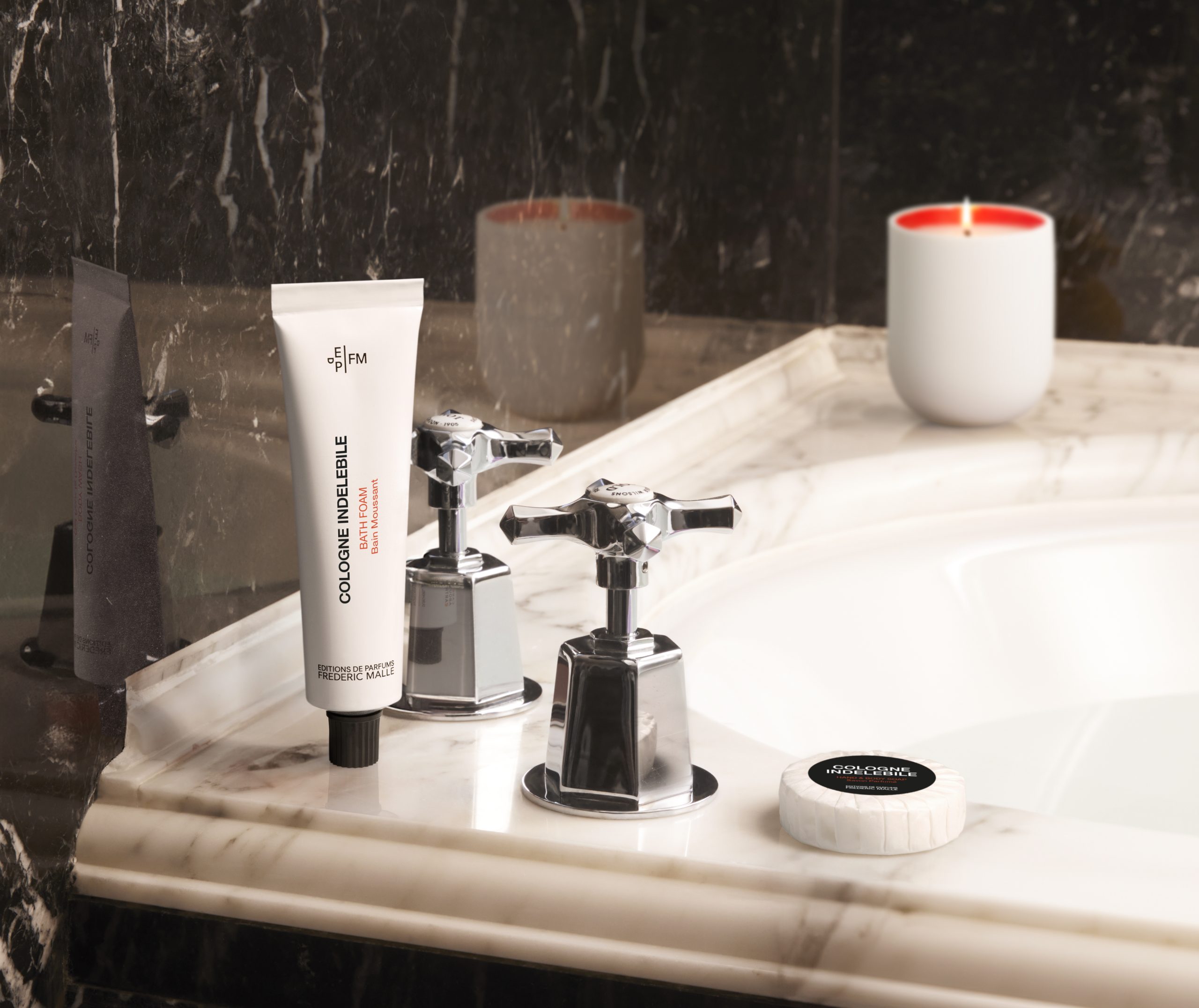 Die Tube mit dem Badeschaum „Cologne Indélébile“ von Editions de Parfums Frédéric Malle steht auf einem Marmorwaschtisch neben verchromten Wasserhahngriffen, zusammen mit einem Stück Seife und einer Kerze in einem eleganten Badezimmer.
