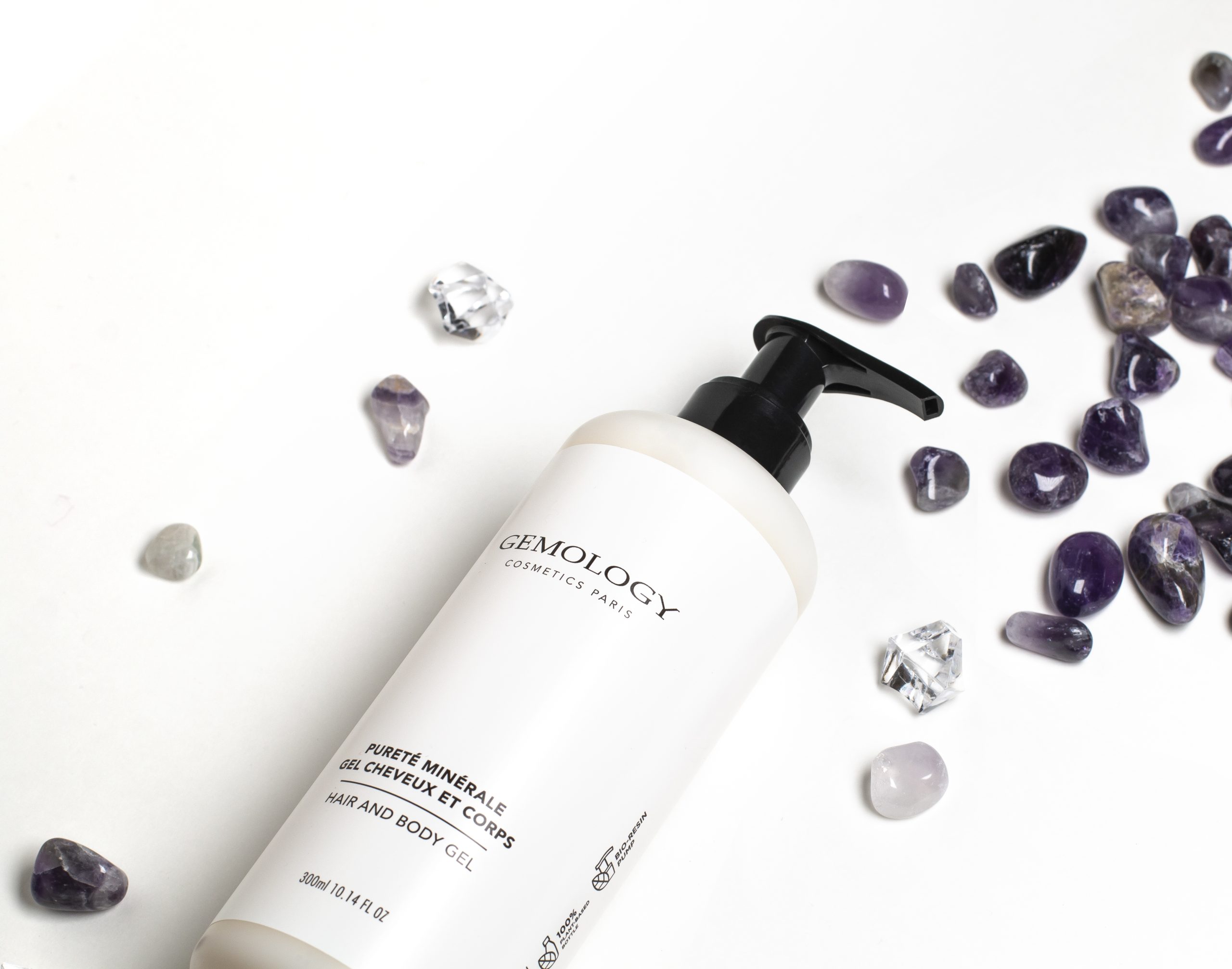 Gemology Kosmetik: Eine Flasche mit Haar- und Körpergel von Paris vor weißem Hintergrund, umgeben von verstreuten violetten Edelsteinen und klaren Kristallstücken, die eine von Mineralien inspirierte Ästhetik unterstreichen.