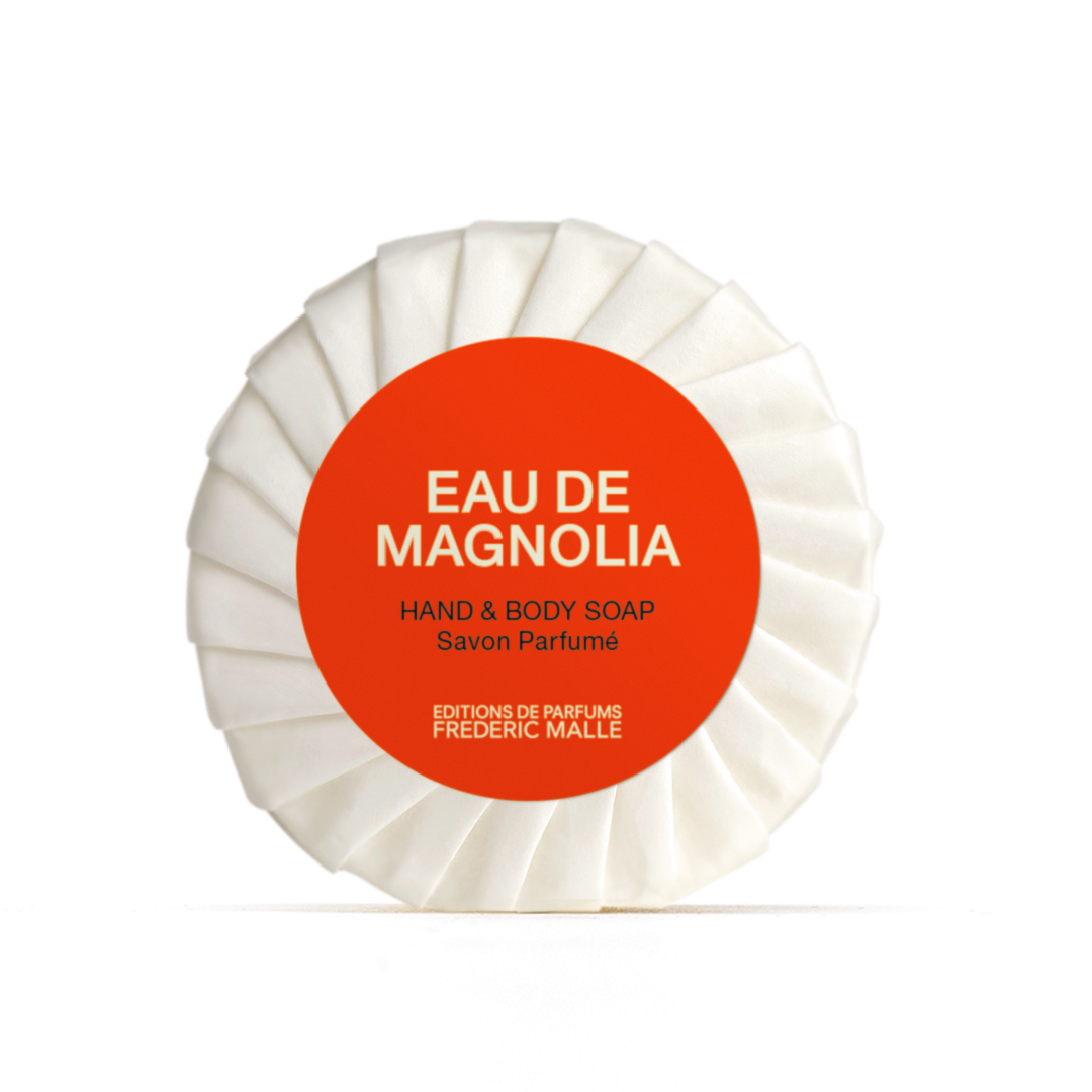 Runde, in Papier eingewickelte „Eau de Magnolia“-Hand- und Körperseife von Editions de Parfums Frédéric Malle mit weißer, plissierter Verpackung und einem leuchtend roten, runden Etikett, das mittig auf einem schlichten, hellen Hintergrund angebracht ist.