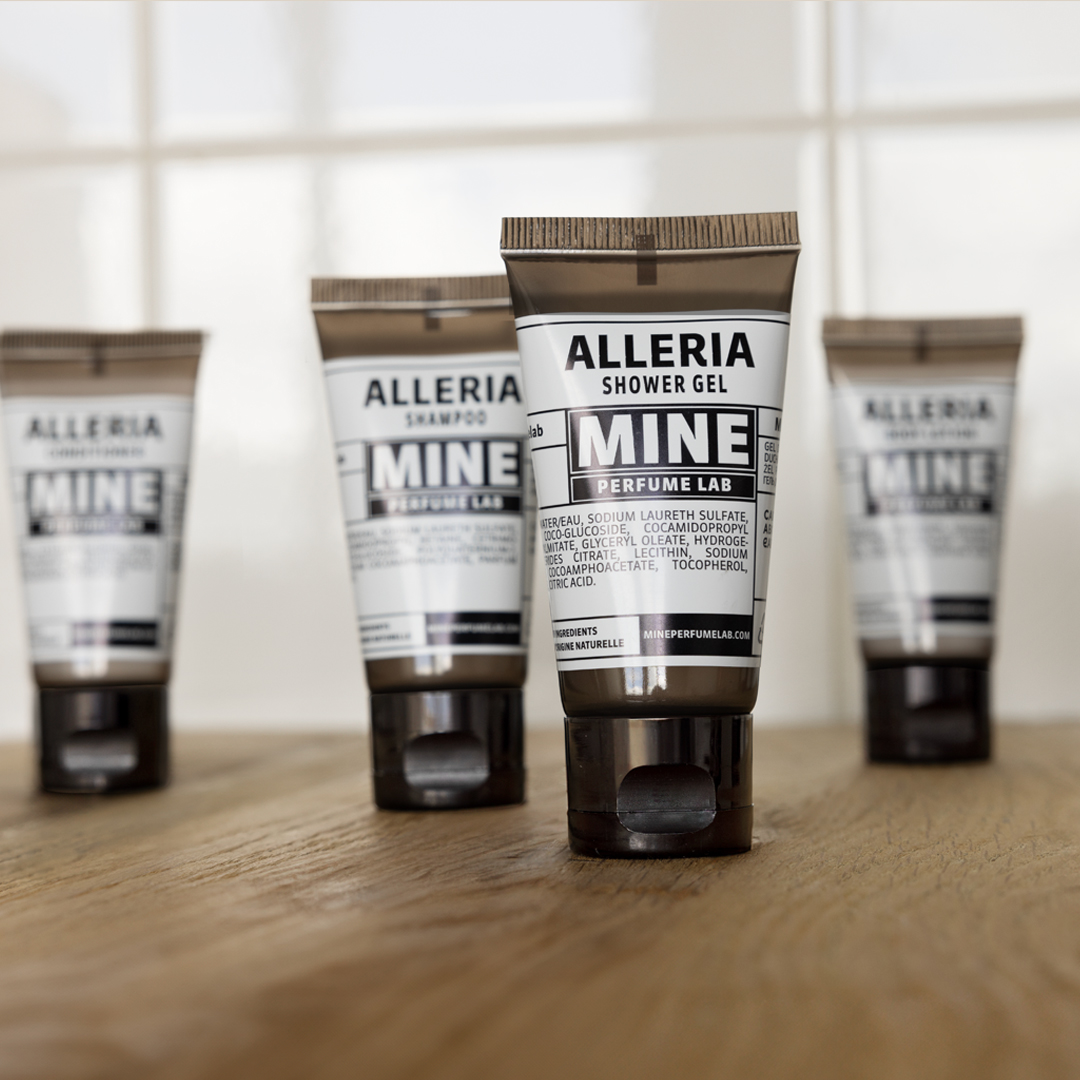 Four Alleria Mine Perfume Lab shower gel and shampoo tubes arranged on a wooden surface, with the centered tube in focus and the others softly blurred against a tiled background.