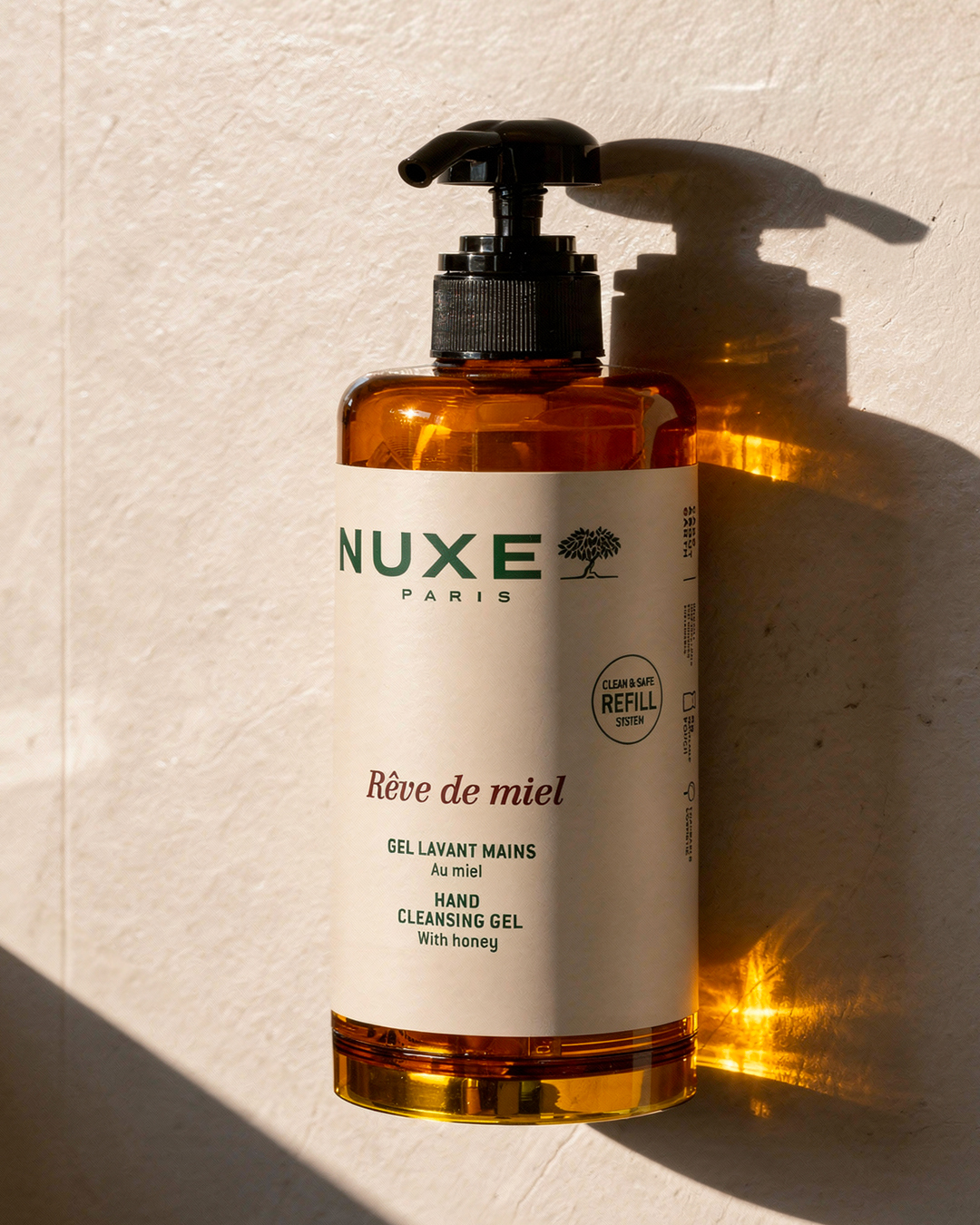 Eine bernsteinfarbene Pumpflasche mit dem Handwaschgel „Rêve de miel“ von NUXE vor einer hellen Wand, auf der das Sonnenlicht goldene Reflexe und einen deutlichen Schatten wirft.