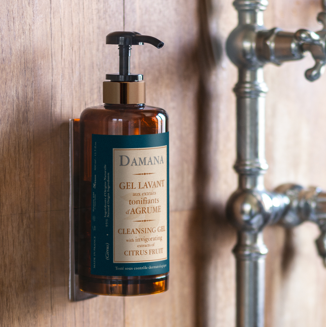 Eine Flasche Damana-Reinigungsgel mit Pumpspender, die an einer Holzwand neben Metallrohren befestigt ist, wobei die Inhaltsstoffe auf Zitrusbasis und eine rustikale Badezimmerkulisse im Vordergrund stehen.