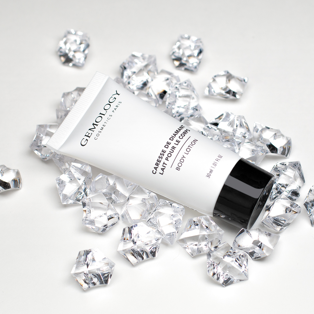 Gemology Kosmetik: Eine Tube Körperlotion von Paris, platziert zwischen verstreuten klaren Kristallsteinen auf weißem Hintergrund, unterstreicht eine klare, von Mineralien inspirierte Ästhetik.