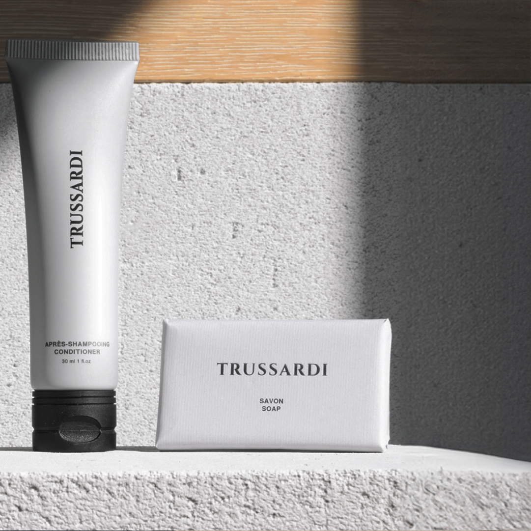 Trussardi Eine aufrecht stehende Spülspülflasche neben einem verpackten Seifenstück auf einer strukturierten Oberfläche, beleuchtet von sanftem Tageslicht, das warme Schatten wirft und das klare, minimalistische Design unterstreicht.