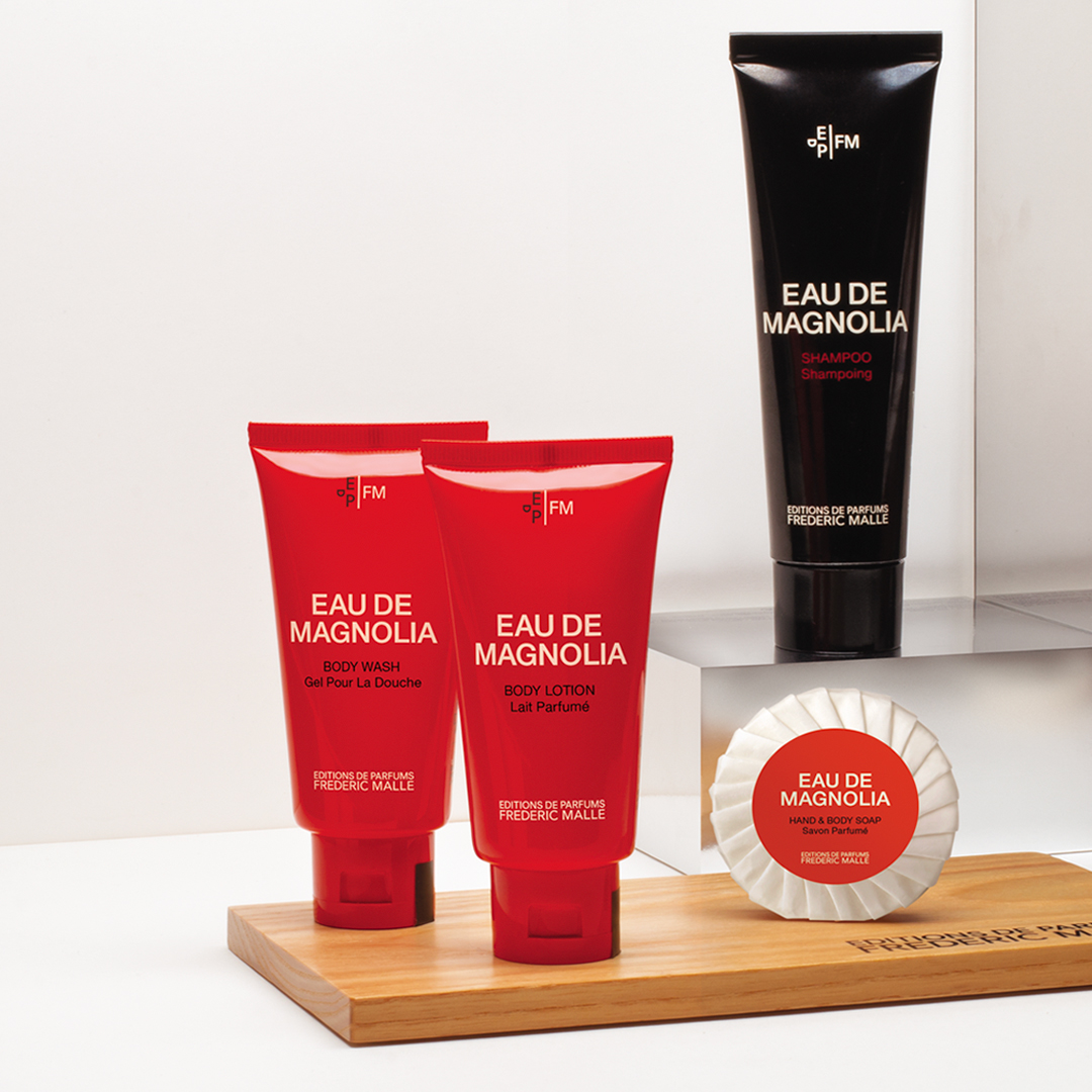Die „Eau de Magnolia“-Badezusätze von Editions de Parfums Frédéric Malle, arrangiert auf einem Holztablett, bestehend aus zwei roten Tuben (Duschgel und Körperlotion), einer schwarzen Shampootube und einer verpackten Seife, präsentiert in einem klaren, minimalistischen Raum.