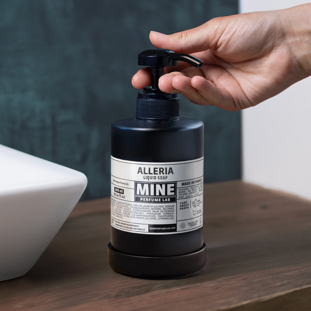 Alleria Mine Perfume Lab liquid soap bottle with a black pump on a wooden countertop next to a white sink, as a hand presses the dispenser, highlighting a modern bathroom setting with a dark textured wall.