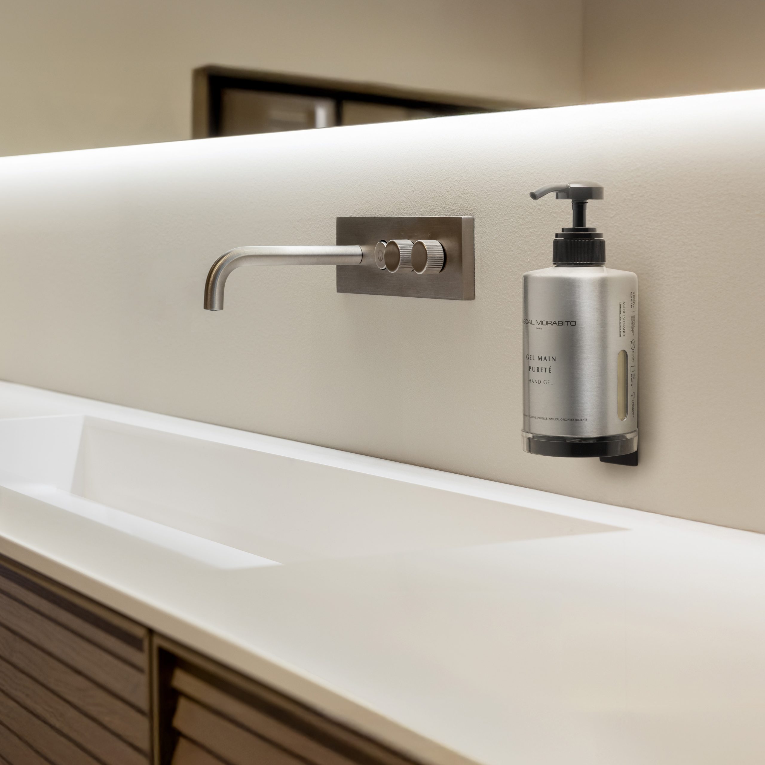 Pascal Morabito Eine Flasche Handdesinfektionsmittel neben einem modernen Waschbecken mit Wasserhahn aus gebürstetem Metall in einem sanft beleuchteten Badezimmer.