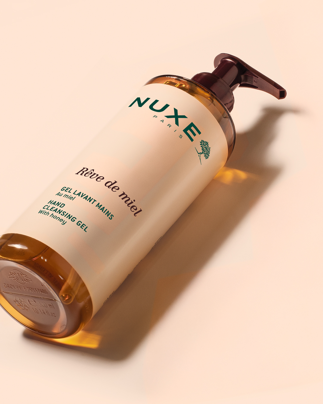 NUXE Rêve de miel Handwaschgel in einer Pumpflasche, die auf einem beigen Hintergrund schräg steht; die bernsteinfarbene Flüssigkeit ist sichtbar, und sanftes Licht wirft elegante Schatten.