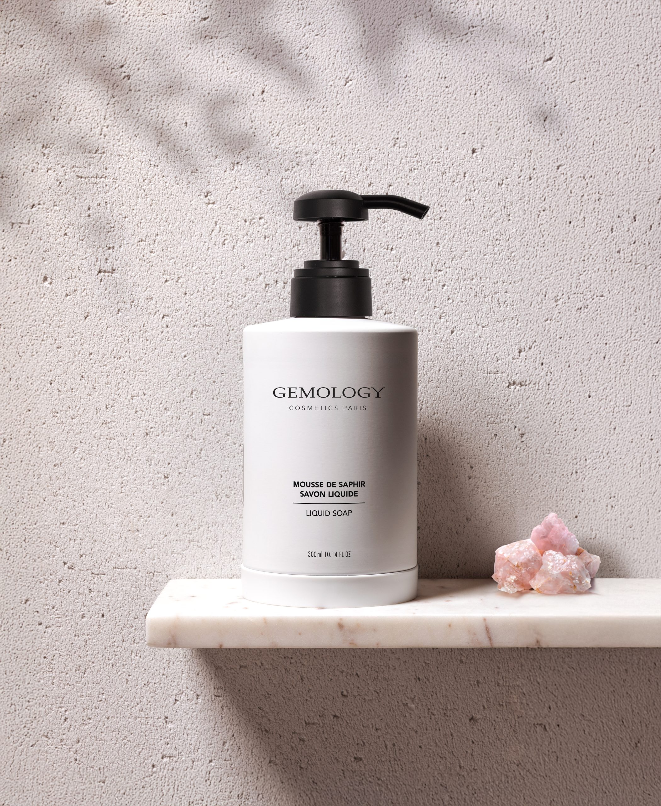 Gemology Eine Flasche Flüssigseife von Cosmetics Paris mit schwarzer Pumpe, präsentiert auf einem hellen Marmorregal vor einer strukturierten beigen Wand, ergänzt durch einen rosa Mineralstein, schafft eine minimalistische und elegante Kulisse.