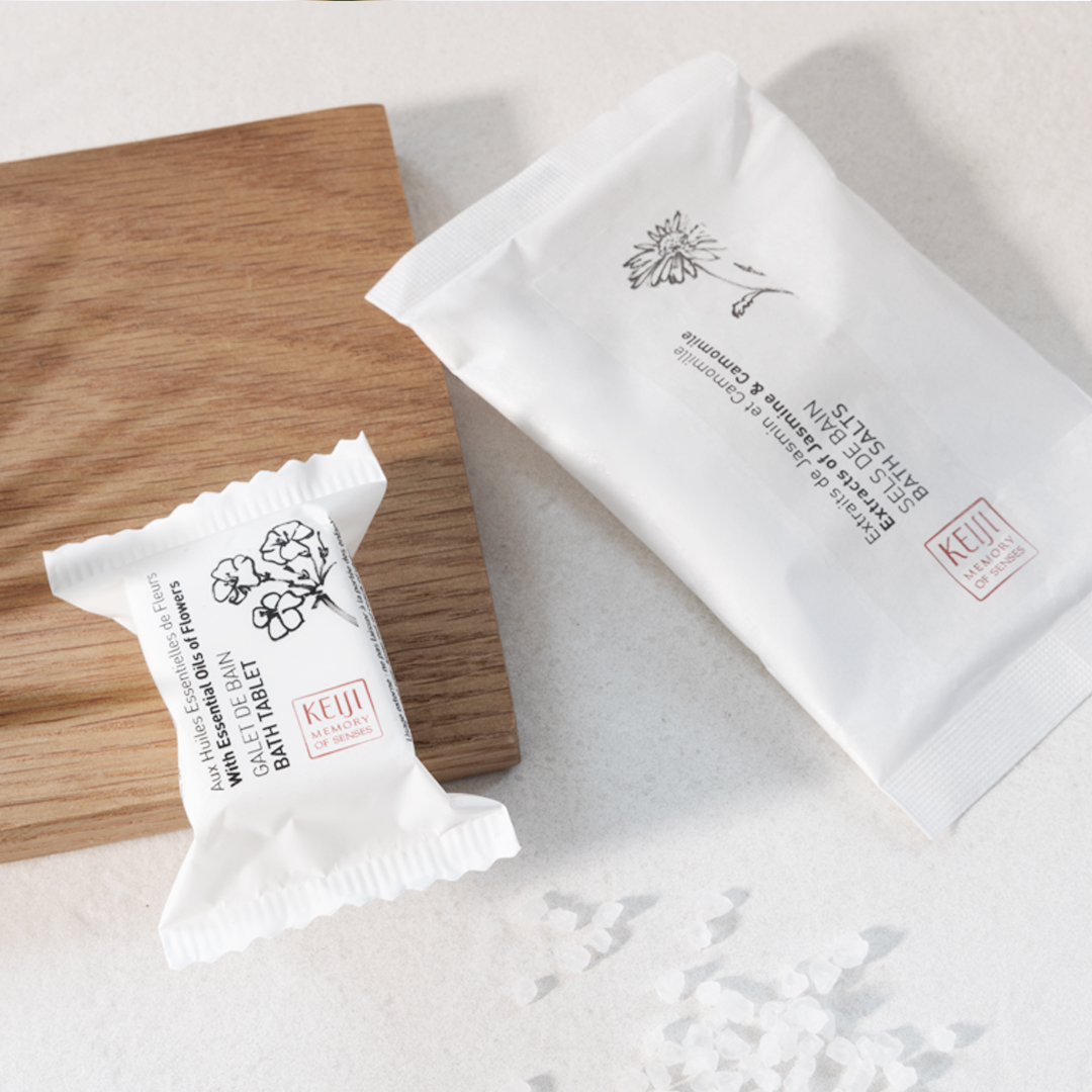Keiji Badezusätze der Marke „Memory of Senses“ auf einer hellen Fläche präsentiert: einzeln verpackte Badetabletten und Badesalz in weißer Verpackung neben einem Holztablett, wobei verstreute Salzkristalle für ein klares, vom Spa inspiriertes Ambiente sorgen.