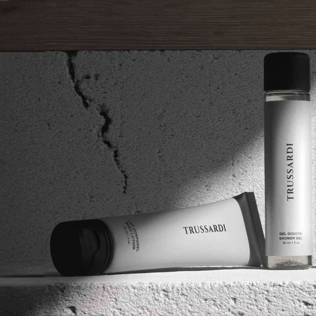 Trussardi Eine Tube Körperlotion und eine Flasche Duschgel von Trussardi, präsentiert auf einem Regal aus strukturiertem Beton, wobei eine dramatische Beleuchtung starke Schatten wirft und die minimalistische schwarz-weiße Verpackung hervorhebt.