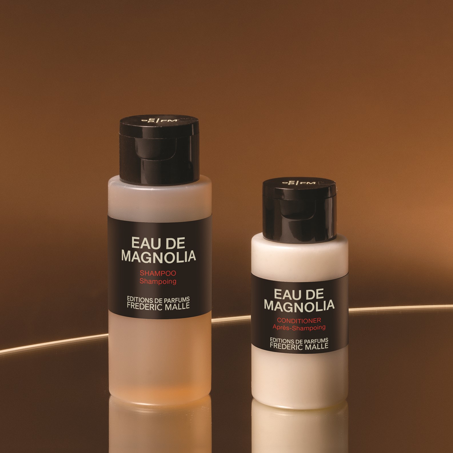 Nahaufnahme der Shampoo- und Spülungsflaschen „Eau de Magnolia“ von Editions de Parfums Frédéric Malle, präsentiert vor einem warmen braunen Hintergrund, wobei sanfte Reflexe die mattierten Flaschen und schwarzen Verschlüsse hervorheben.