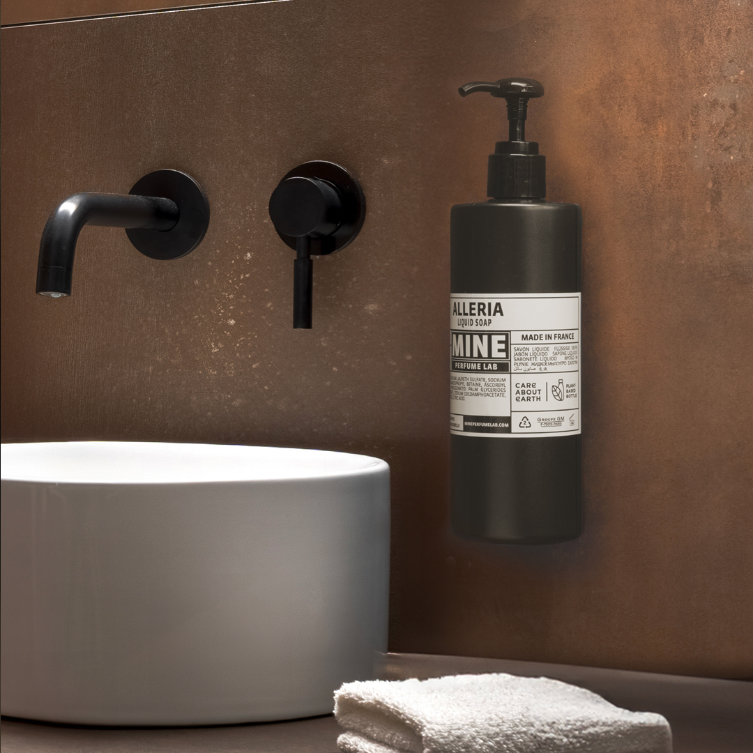 Wall-mounted Alleria Mine Perfume Lab liquid soap bottle with a black pump next to a modern white sink, set against a warm brown textured wall with minimalist black fixtures.