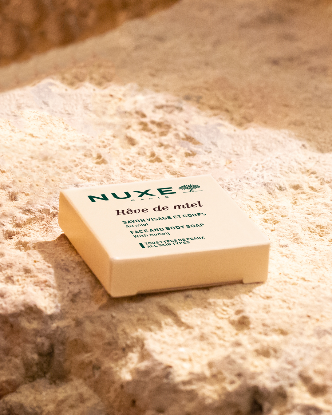 Die NUXE Rêve de miel Gesichts- und Körperseife liegt auf einer strukturierten, sandartigen Oberfläche und wird von warmem, natürlichem Licht in Szene gesetzt, das die beigefarbene Verpackung und den grünen Schriftzug zur Geltung bringt.