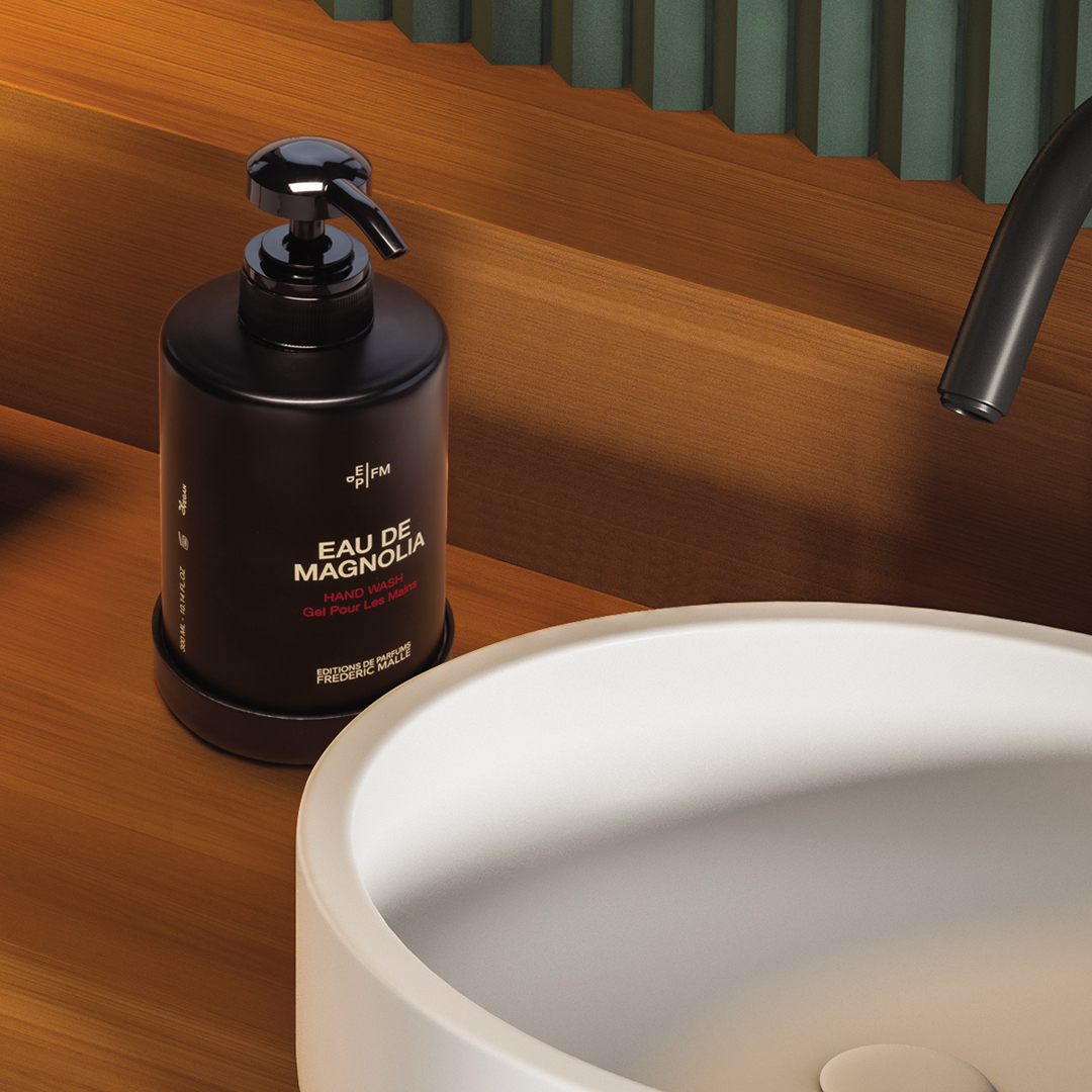 Eine Flasche „Eau de Magnolia“-Handseife mit schwarzer Pumpe steht neben einem modernen weißen Waschbecken auf einer warmen Holzarbeitsplatte, im Hintergrund sind ein geschwungener schwarzer Wasserhahn und eine strukturierte grüne Wand zu sehen.