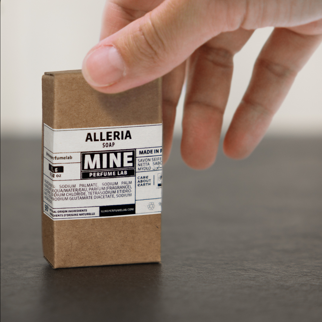 Hand placing a boxed Alleria Mine Perfume Lab soap on a dark surface, with minimalist brown packaging and a technical-style label in black and white.