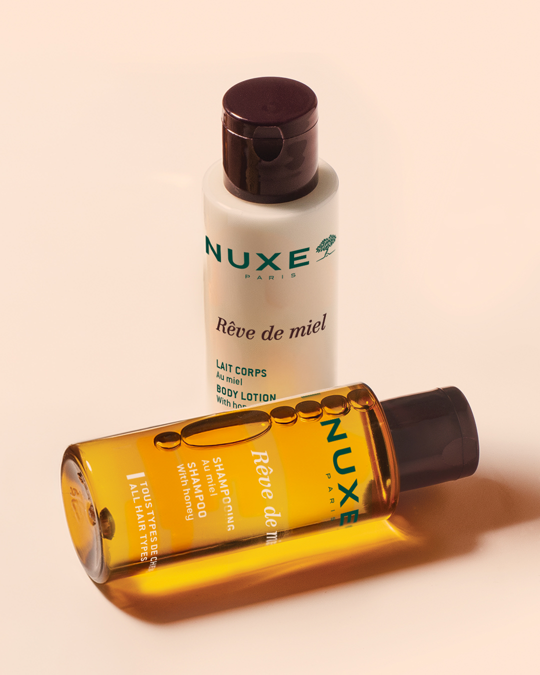 Produkte der Serie „Rêve de miel“ von NUXE Paris vor beigem Hintergrund: eine aufrecht stehende Körperlotion in Honigfarbe und eine horizontal liegende, bernsteinfarbene Shampooflasche mit warmen Lichtreflexen.
