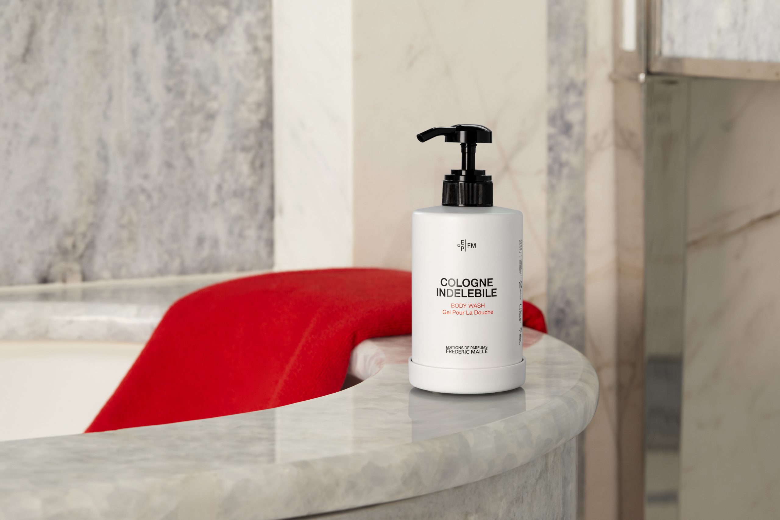 Eine Flasche „Cologne Indélébile“-Duschgel steht auf einer Marmorplatte neben einer Badewanne, im Hintergrund liegt ein rotes Handtuch – so entsteht eine warme und elegante Badezimmeratmosphäre.