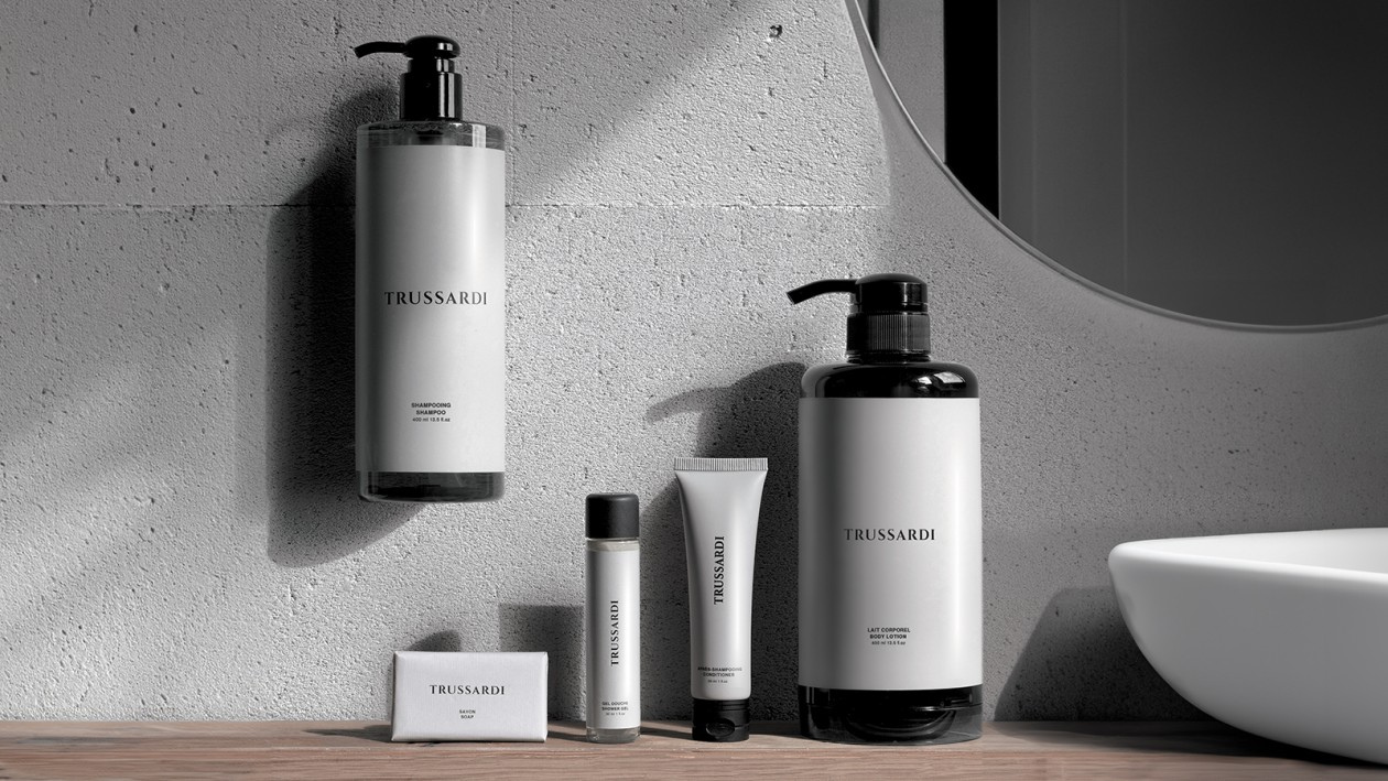 Minimalist bir banyo ortamında ahşap bir tezgahın üzerinde sergilenen Trussardi ve vücut bakım ürünleri serisi; beyaz bir lavabo ve yuvarlak aynanın yanında duvara monte edilmiş bir şampuan şişesi, vücut losyonu pompası, küçük tüpler ve kutulu bir sabun yer alıyor.