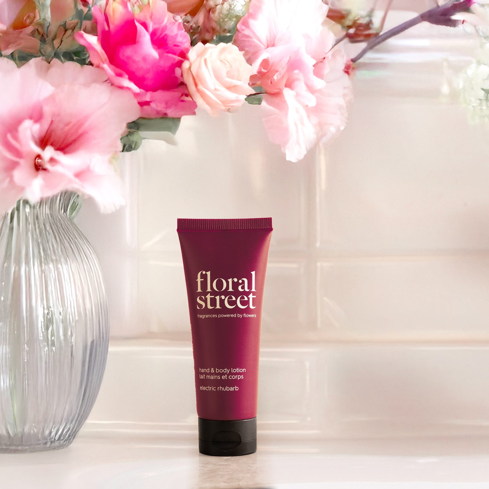 Floral Street Loção para as mãos e para o corpo «Electric Rhubarb» bisnaga ao lado de um vaso de vidro com flores cor-de-rosa e brancas, num ambiente interior suave e luminoso.