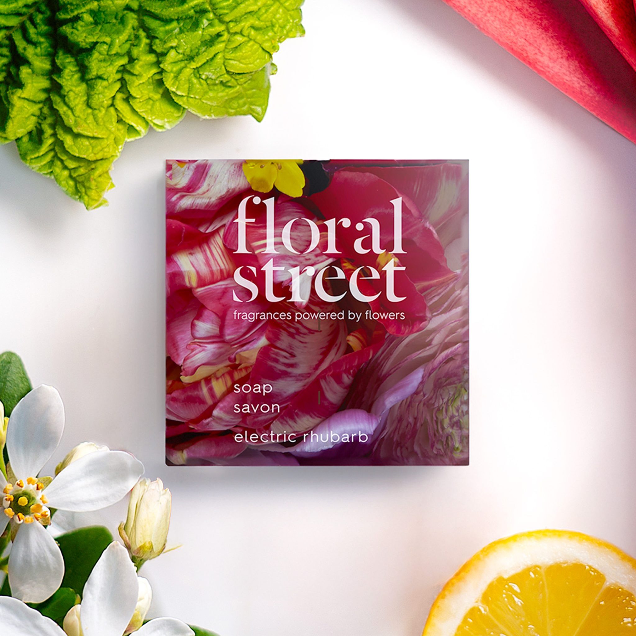 Floral Street Embalagem sabonete «Electric Rhubarb», rodeada por talos de ruibarbo frescos, flores brancas, folhas verdes e uma fatia de limão sobre um fundo branco imaculado.