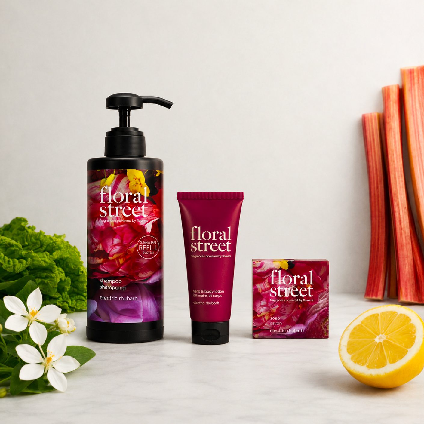 Floral Street Produtos de banho e cuidados corporais da Electric Rhubarb, incluindo champô recarregável, loção para as mãos e para o corpo e sabonete, apresentados com talos de ruibarbo frescos, flores brancas, folhas verdes e limão num fundo simples.